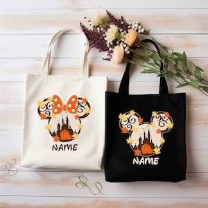 May include: Two tote bags with a Mickey Mouse silhouette design. The silhouette features a castle, pumpkin, and fall leaves. One bag is white with a red bow and the other is black. Both bags have the text "NAME" printed on them.