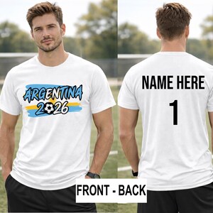 May include: White t-shirt with "ARGENTINA 2026" in blue and black text, a soccer ball graphic, and blue and yellow stripes. The back of the shirt has "NAME HERE" and the number "1" in black.