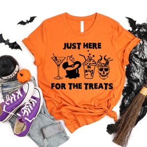 Puede incluir: Camiseta naranja con el texto "JUST HERE FOR THE TREATS" y gráficos de Halloween. El diseño incluye un martini, un sombrero de bruja, una bebida y una calavera. La imagen incluye zapatillas moradas, vaqueros y decoraciones de Halloween.