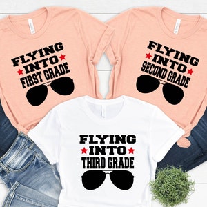 Puede incluir: Tres camisetas de color melocotón y blanco con el texto "Flying into" y el nivel de grado, "First Grade", "Second Grade" y "Third Grade". Cada camiseta tiene un par de gafas de sol y una estrella roja a cada lado de la palabra "into".