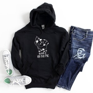 Puede incluir: Sudadera con capucha negra con un cerdo de dibujos animados blanco y el texto "Blame it on the pig".