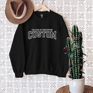 May include: Black sweatshirt with white text that reads "College of Education Custom" hanging on a hanger against a white background with a brown hat and a cactus with Christmas lights.