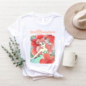 A white t-shirt with a Disney-themed graphic, a beige hat, a mug, and some greenery on a white surface.