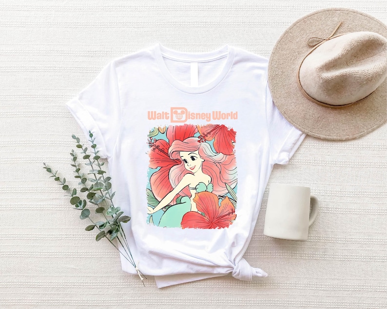 A white t-shirt with a Disney-themed graphic, a beige hat, a mug, and some greenery on a white surface.