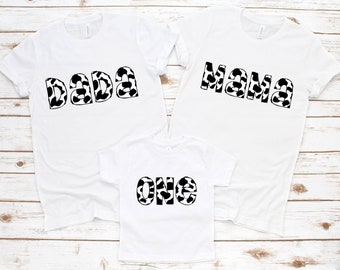 Könnte beinhalten: Drei weiße T-Shirts mit schwarz-weißem Kuhmuster-Schriftzug. Die Shirts sagen "Dada", "Mama" und "Eins".