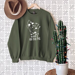 Puede incluir: Una sudadera verde con un cerdo de dibujos animados blanco y el texto "Blame it on the pig".