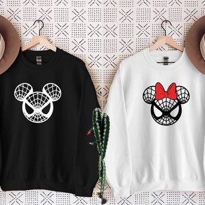 May include: Two black and white sweatshirts with a Spider-Man Mickey Mouse design. One sweatshirt is black with a white design and the other is white with a black design. The white sweatshirt has a red bow on top of the head.