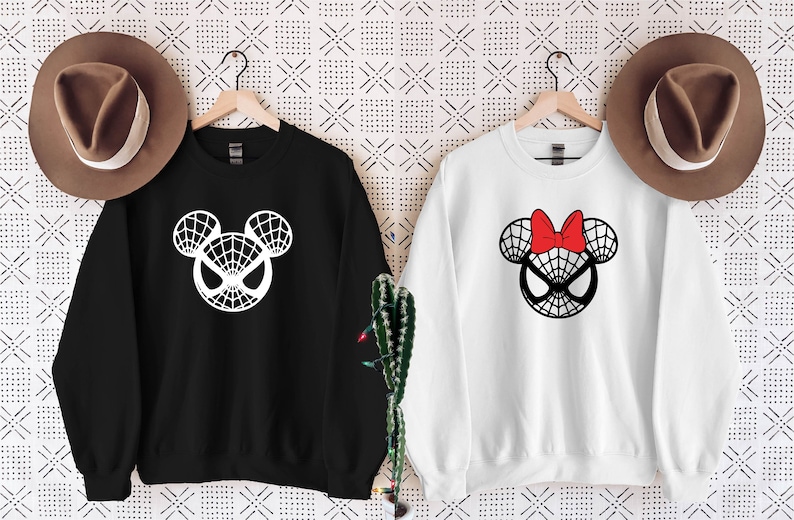 May include: Two black and white sweatshirts with a Spider-Man Mickey Mouse design. One sweatshirt is black with a white design and the other is white with a black design. The white sweatshirt has a red bow on top of the head.
