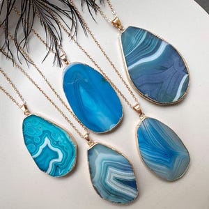 Op de afbeelding: Vijf gouden kettingen met blauwe agaat hangers. Elke hanger heeft een andere blauwe tint en een uniek patroon van witte lijnen.