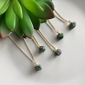 Peut inclure: Cinq colliers en or avec des pendentifs en pierres précieuses vertes et noires. Les colliers sont disposés en éventail sur une surface blanche.
