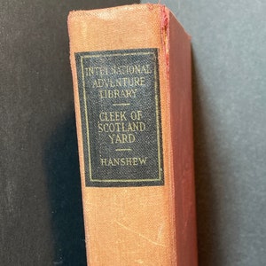 May include: A vintage book with a red spine and a tan cover. The title is "International Adventure Library" and the book is titled "Cleek of Scotland Yard" by "Hanshew".
