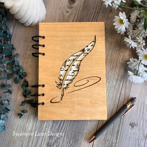 May include: A wooden journal with a spiral binding features a laser-cut design of a quill pen with a feather made of script. The journal is on a woodgrain surface.