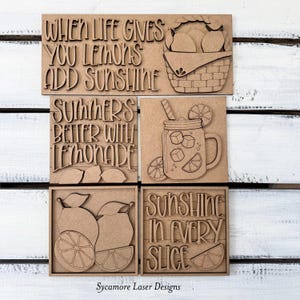 May include: A set of four wooden signs with laser-cut designs and text. The signs feature phrases like "When life gives you lemons, add sunshine" and images of lemons, lemonade, and a basket. The signs are on a white wooden surface.