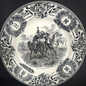 May include: Black and white ceramic plate with a decorative border and a scene of two men on horseback. The scene depicts a battle with soldiers in the background. The text "SORTIE DE DESAIX A MARENGO" is printed on the plate.