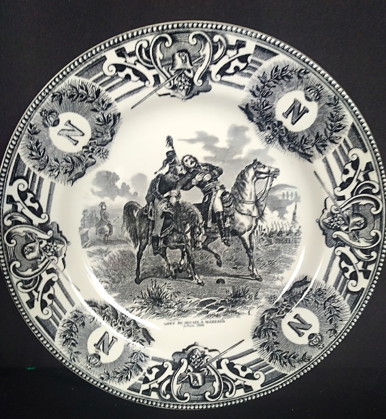 May include: Black and white ceramic plate with a decorative border and a scene of two men on horseback. The scene depicts a battle with soldiers in the background. The text "SORTIE DE DESAIX A MARENGO" is printed on the plate.