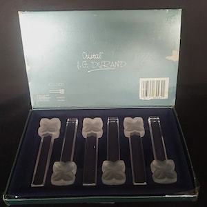Puede incluir: Un juego de seis anillos de servilleta de cristal transparente en una caja azul. La caja tiene las palabras "Cristal" y "J.G. DURAND" impresas.