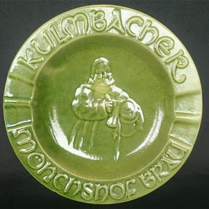Könnte beinhalten: Grüner Aschenbecher aus Keramik mit einem Reliefbild einer Person, die eine Tasse hält. Die Worte "Kulmbacher Munchner Brau" sind um den Rand des Aschenbechers geschrieben.
