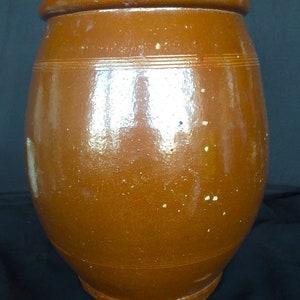 May include: A large, brown, glazed ceramic crock with a rounded body and a wide opening. The crock has a slightly textured surface and appears to be antique.