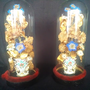 May include: Two glass domes with gold-painted flowers and blue accents in white porcelain vases. The flowers are arranged in a bouquet and are displayed on a black base with a red velvet trim.