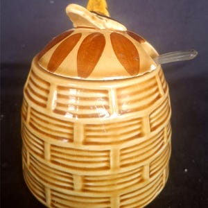 Op de afbeelding: Een keramische honingpot met deksel en een kleine lepel. De pot is bruin met een mandweefpatroon. Het deksel is bruin met bruine ovale accenten en een gele bijvormige handgreep. Een doorzichtige lepel rust op het deksel.