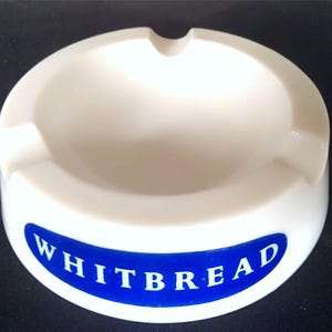 May include: White ceramic ashtray with a blue oval label that reads "WHITBREAD".