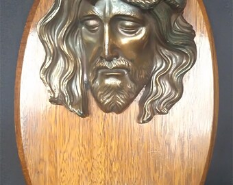 Könnte beinhalten: Eine ovale Holzplakette mit einer Bronzeskulptur von Jesu Gesicht mit Dornenkrone. Ein kleines Bronzebecken ist an der Unterseite der Plakette befestigt.