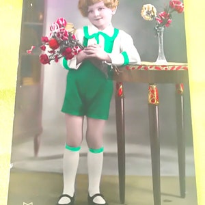 緑のパンツスーツと白いシャツを着た素敵な子供が、花瓶と花束を手にアールデコ調のテーブルに立っています。 1930年代