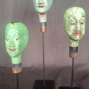 Op de afbeelding: Drie houten poppenkoppen met geschilderde gezichten op zwarte metalen standaards. De koppen zijn groen en hebben overdreven kenmerken.