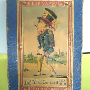 Op de afbeelding: Een vintage doos voor garen, met een cartoonillustratie van een man met een hoge hoed en een wandelstok. De doos is blauw met gouden versieringen en de tekst "Fil en Capsule" en "50 Meter Garantie".