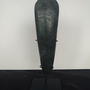 May include: A dark green, elongated stone axe head with a rounded top and a tapered bottom. The axe head is mounted on a black metal stand. The stone has a textured surface with visible cracks.