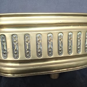 May include: A brass planter with a decorative design and two handles. The planter is oval shaped and has a raised base.