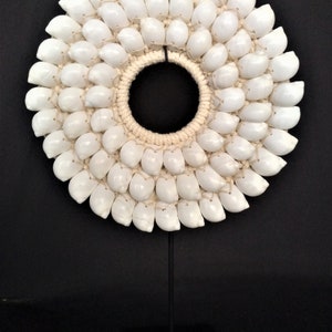 Op de afbeelding: Een witte schelpenketting met een geweven koord, gepresenteerd op een zwarte standaard. De ketting is gemaakt van veel kleine, witte schelpen die in een cirkelvormig patroon zijn gerangschikt.
