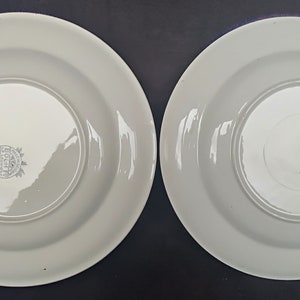 May include: Two white ceramic plates with a raised rim. The plates have a simple design and a small logo on the bottom.