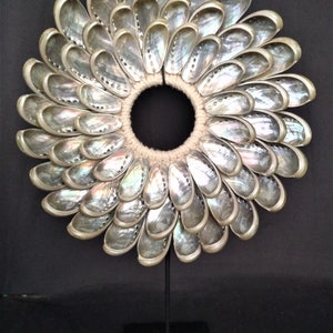 Könnte beinhalten: Eine runde Halskette aus schillernden weißen Muscheln, die in einem blumenartigen Muster angeordnet sind, auf einem schwarzen Ständer.