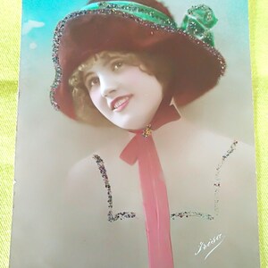 Jolie jeune femme portant un chapeau marron avec un nœud vert et un nœud rose autour du cou. Carte de la marque Frisa. Traces de paillettes sur la carte.