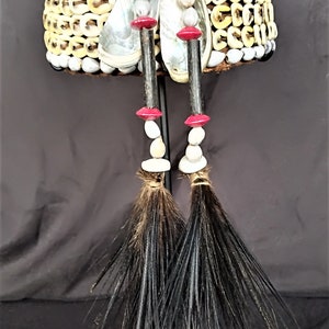 Peut inclure: Une coiffe traditionnelle faite de coquillages tissés, avec deux longues touffes de plumes noires qui pendent du bas. La coiffe est décorée de perles blanches et rouges.