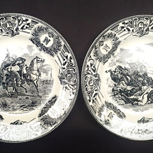 May include: Two black and white porcelain plates with decorative borders and images of historical battles. The plates feature the letter "N" in a decorative wreath.