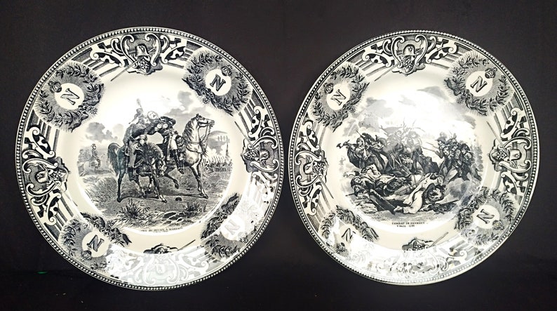 May include: Two black and white porcelain plates with decorative borders and images of historical battles. The plates feature the letter "N" in a decorative wreath.