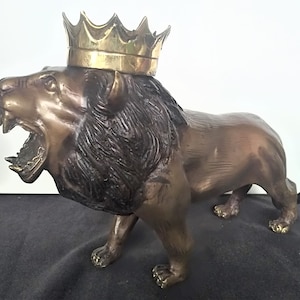 Könnte beinhalten: Eine Bronzelöwenstatue mit einer goldenen Krone auf dem Kopf. Der Löwe brüllt mit offenem Maul und seine Mähne ist detailliert. Die Statue steht auf einer schwarzen Oberfläche.
