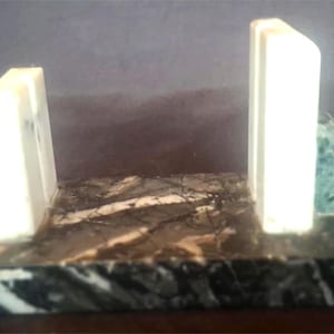 Op de afbeelding: Marmeren boekensteunen met een rechthoekige basis en twee staande steunen. De basis is een mix van zwart, wit en grijs, terwijl de steunen wit zijn met groene driehoekige accenten. Een decoratief item.