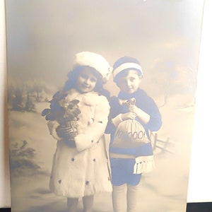 Cartão de Ano Novo Alemão da década de 1920, Duas crianças com casacos de pele e lindos chapéus, A menina com um arranjo de flores e o menino com uma bolsa