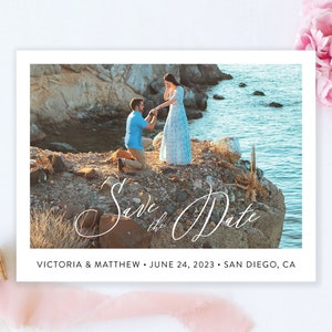 Può includere: Carta "Save the Date" con una foto di una coppia su una scogliera rocciosa che si affaccia sull'oceano. Il testo sulla carta recita "Save the Date" in un carattere corsivo e "Victoria & Matthew - 24 giugno 2023 - San Diego, CA".
