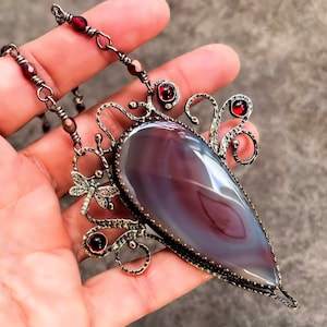 Puede incluir: Un collar de plata con un gran colgante de ágata en forma de lágrima. El colgante está rodeado de intrincados trabajos de alambre de plata y acentuado con pequeñas cuentas de vidrio rojas. El collar está en una mano.