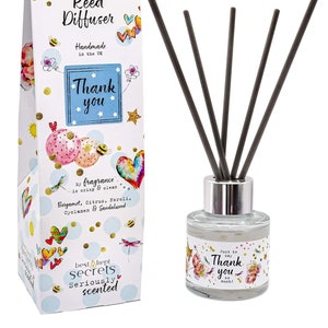 Pode incluir: Uma caixa branca com um design colorido que apresenta corações, estrelas e abelhas. A caixa diz "Just Because... Reed Diffuser" e "Thank you". A caixa está ao lado de uma garrafa de vidro transparente com uma tampa prateada e varas pretas. A garrafa tem um design colorido com flores e o texto "Just to say Thank you so much!"