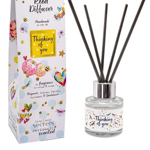 May include: A white box with a colorful design and the text "Thinking of you" and "Just Because... Reed Diffuser" with a clear glass bottle with a silver lid and black reeds. The bottle has a label with the text "Thinking of you" and a colorful design.