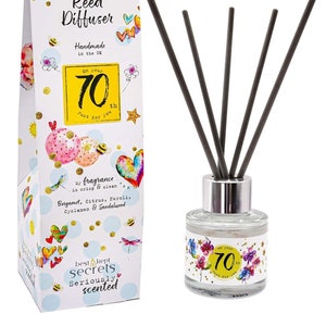 May include: A white box with colourful illustrations and the text "Just Because... Reed Diffuser Handmade in the UK On your 70th Just for you" and the text "best kept secrets Seriously scented". A clear glass bottle with a silver lid and black reeds. The bottle has a colourful illustration and the text "On your 70th Just for you".