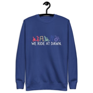 Könnte beinhalten: Blaues Sweatshirt mit dem Text "WE RIDE AT DAWN." Der Text befindet sich unter einer Reihe farbenfroher Illustrationen von Mobilitätshilfen, darunter ein Roller, ein Rollator und ein Rollstuhl. Das Sweatshirt hängt an einem schwarzen Kleiderbügel.