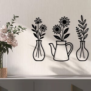 May include: A display of wall art featuring black metal cutouts of flowers in vases and a teapot. A bouquet of pink roses in a clear vase and a decorative box are on a gray surface.