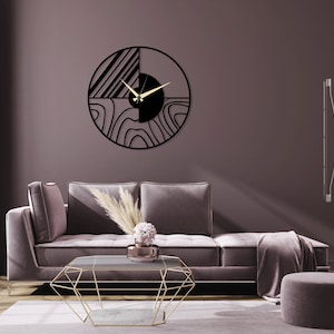 May include: A black and white geometric wall clock with a gold hand, hanging on a mauve wall above a gray sofa with a gold geometric coffee table and a gray ottoman.