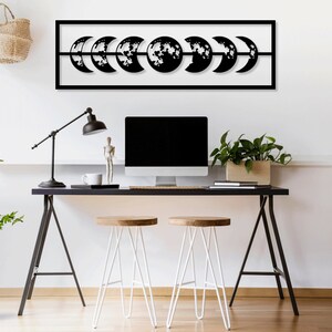 May include: Black metal wall art depicting the phases of the moon in a rectangular frame. The art is hung on a white wall above a black desk with a computer monitor, a lamp, and two white stools.